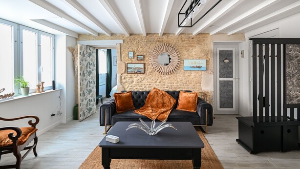 Un salon moderne avec un mur en pierre, un canapé noir agrémenté de coussins orange, une table basse ornée d’un élément décoratif central et un grand miroir.