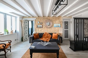 Een moderne woonkamer met een stenen muur, een zwarte bank met oranje kussens, een bijzettafeltje met een decoratief object en een grote spiegel.