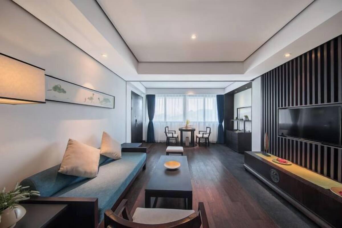 A modern living room with a sofa, a dining table, and chairs. There is a TV on the wall and a window with curtains.