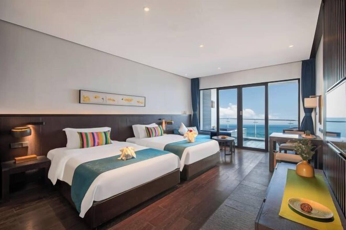 A modern hotel room with two beds, a large window, and a view of the ocean.