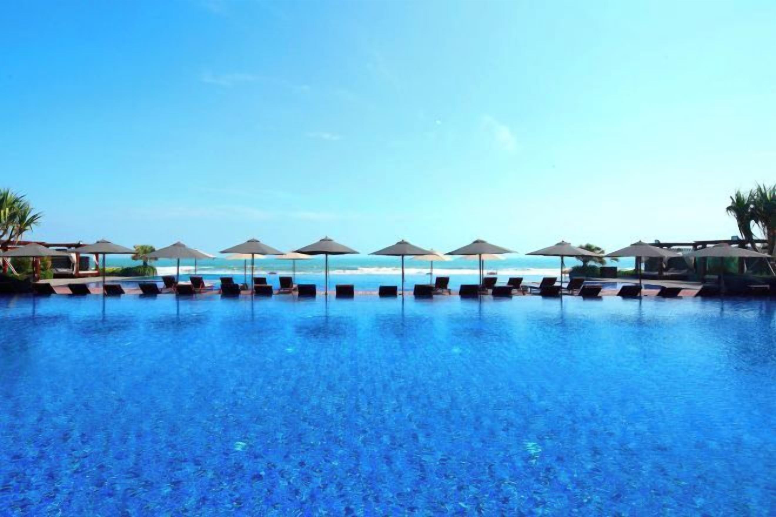 A swimming pool with lounge chairs and umbrellas, a beach in the background, and clear skies.