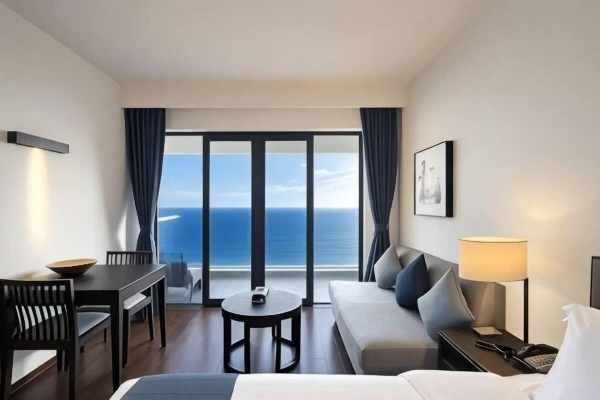 A hotel room with a balcony, a sofa, a dining table, and a lamp.
