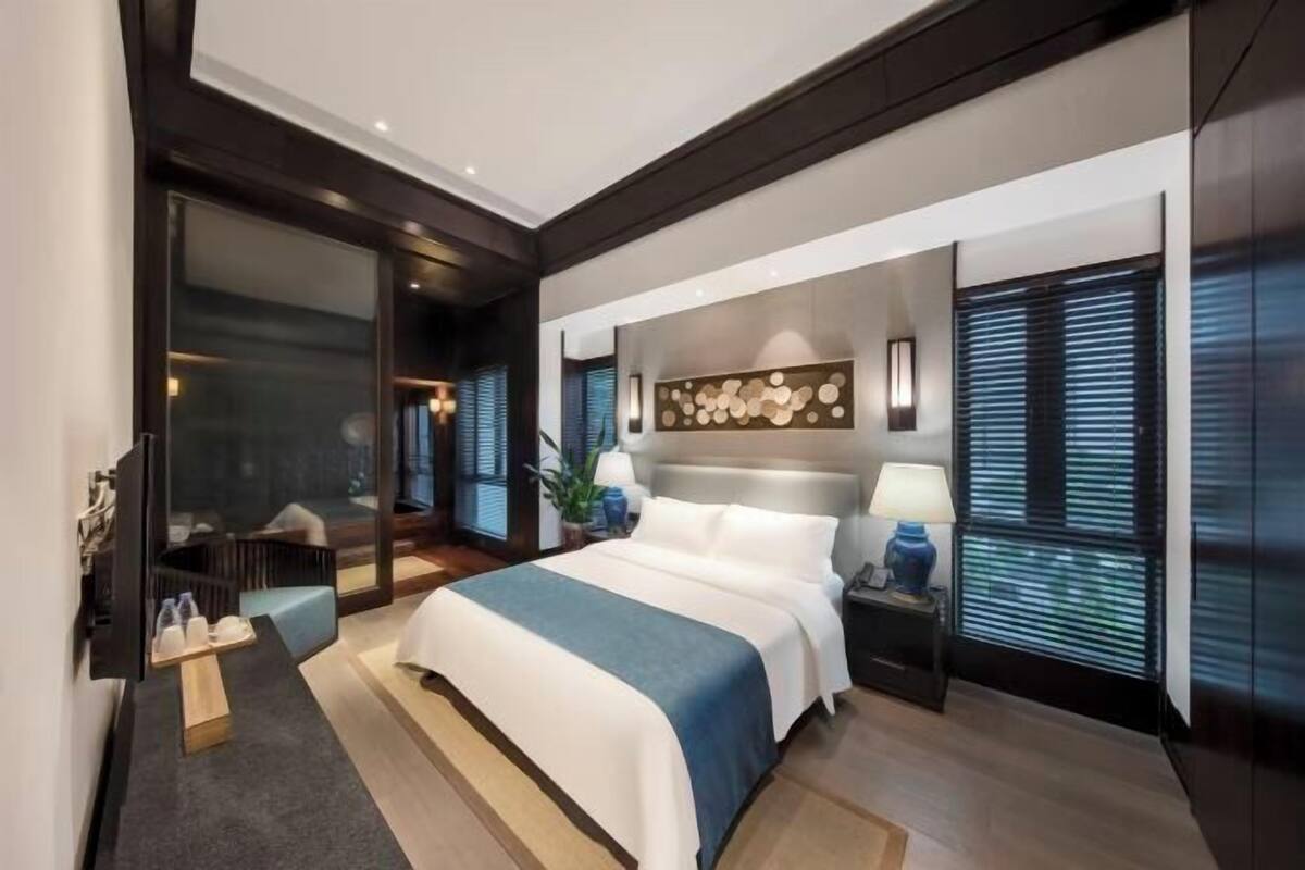 A modern hotel room with a large bed, a bedside table with a lamp, a wall-mounted artwork, and a sliding glass door leading to a balcony.