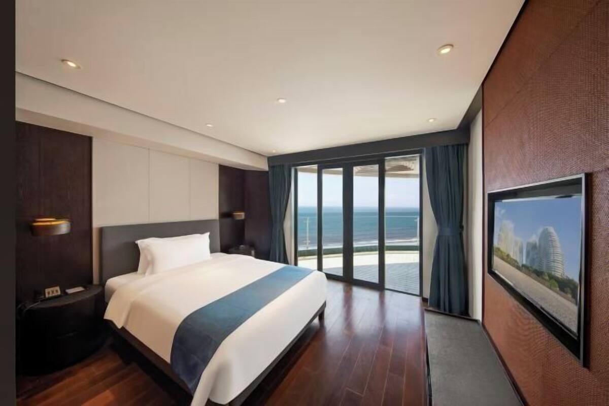 A modern hotel room with a large bed, a nightstand, a wall-mounted TV, and a view of the ocean.