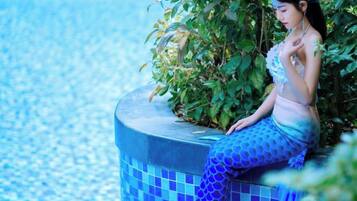 Eine Person sitzt auf einer blauen, gewölbten Struktur mit Fliesen am Pool, im Hintergrund Grünpflanzen.