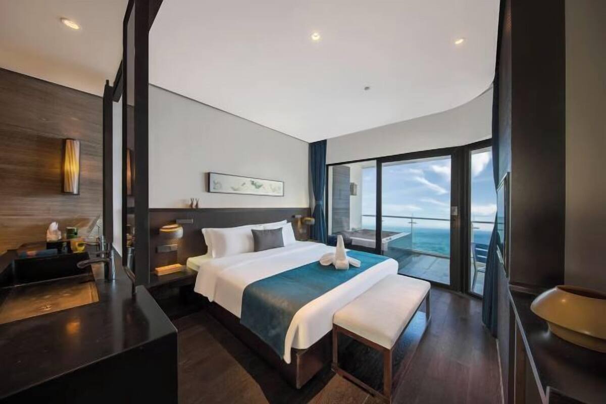 A modern hotel room with a large bed, a bedside table, a bench, a view of the ocean, and a balcony.