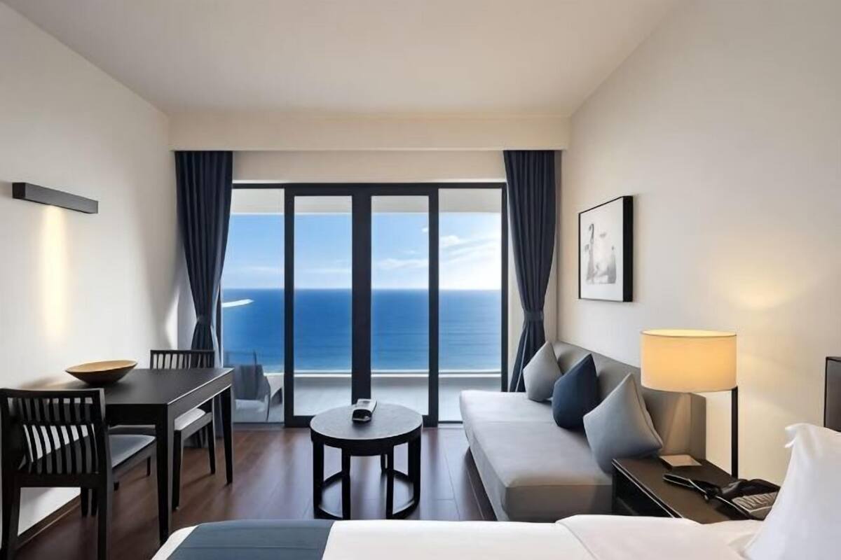 A hotel room with a balcony, a sofa, a dining table, and a lamp.