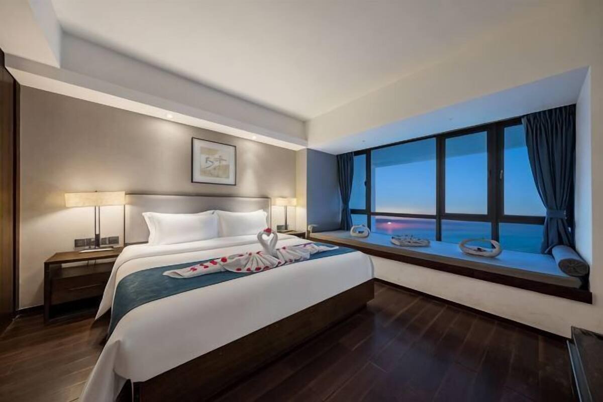 A hotel room with a large bed, two bedside tables with lamps, a window with a view of the sea, and a framed picture on the wall.