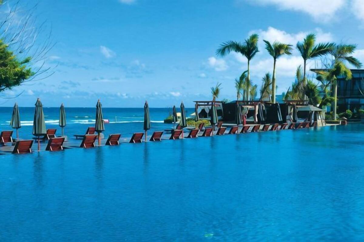 A pool with lounge chairs and umbrellas, palm trees, and a view of the ocean.