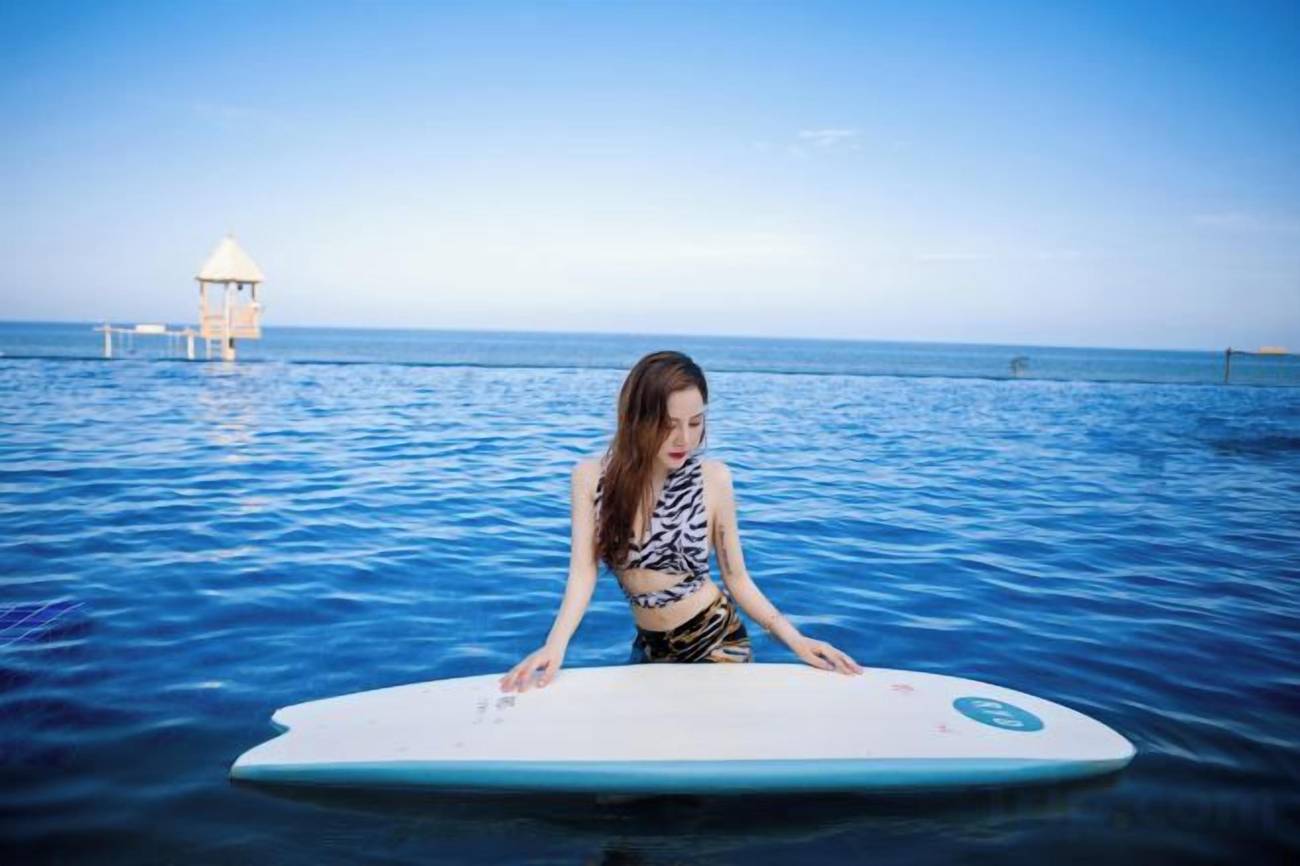 Uma pessoa sentada em uma prancha de surf na água, com um farol ao fundo.