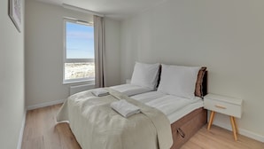 A bedroom with a bed, a nightstand, a window with a view of the beach, and a wooden floor. - Horizon 122 -SeaView by GrandApartments  (Gdansk)