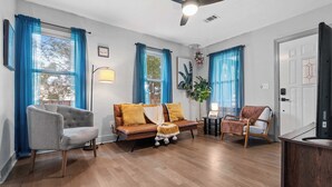 Una sala de estar con sofá, sillón y una mesa de centro. Hay un ventilador de techo, una puerta y una ventana con cortinas.