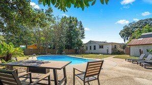 Un patio trasero con piscina, una mesa de madera y sillas de descanso.