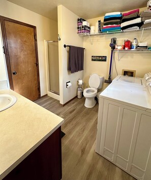 Un bagno con WC, lavandino, doccia e un angolo lavanderia con asciugamani impilati e articoli da toilette.