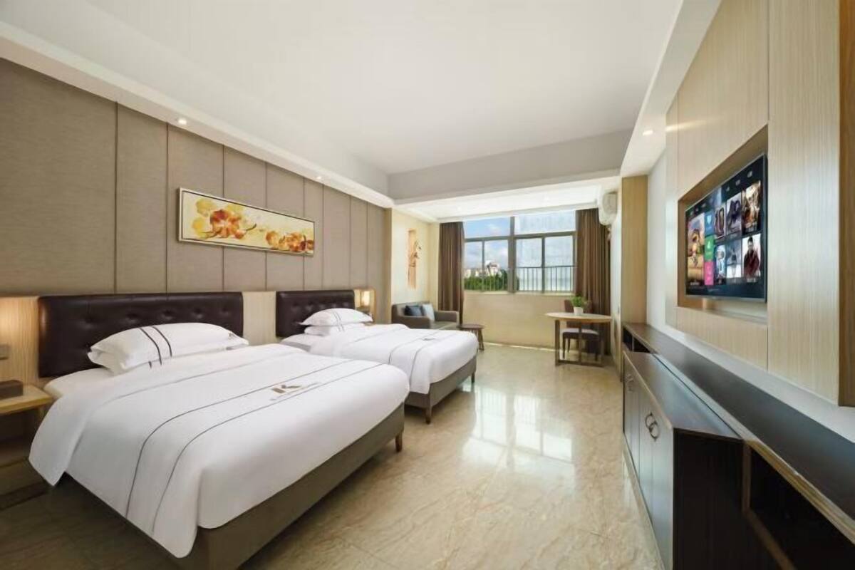 A hotel room with two beds, a TV, a desk, and a window with a view.