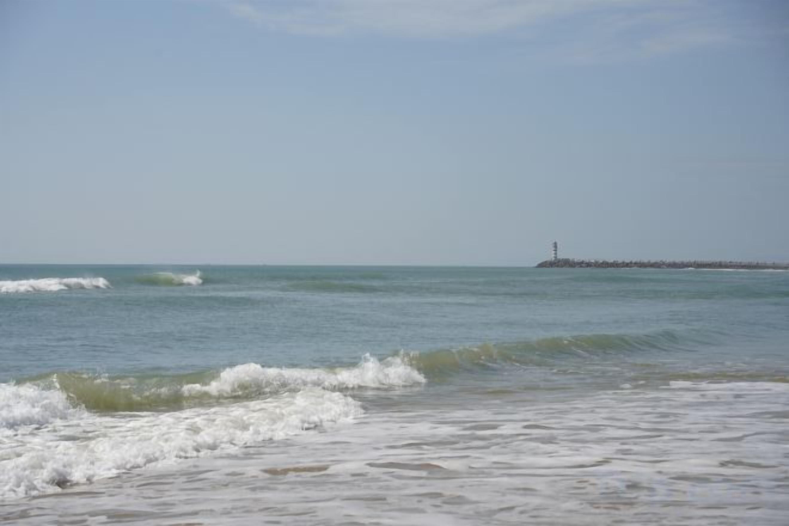 A beach with waves, a pier, and a lighthouse in the distance.