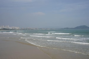 A beach with waves, a distant cityscape, and a mountain.