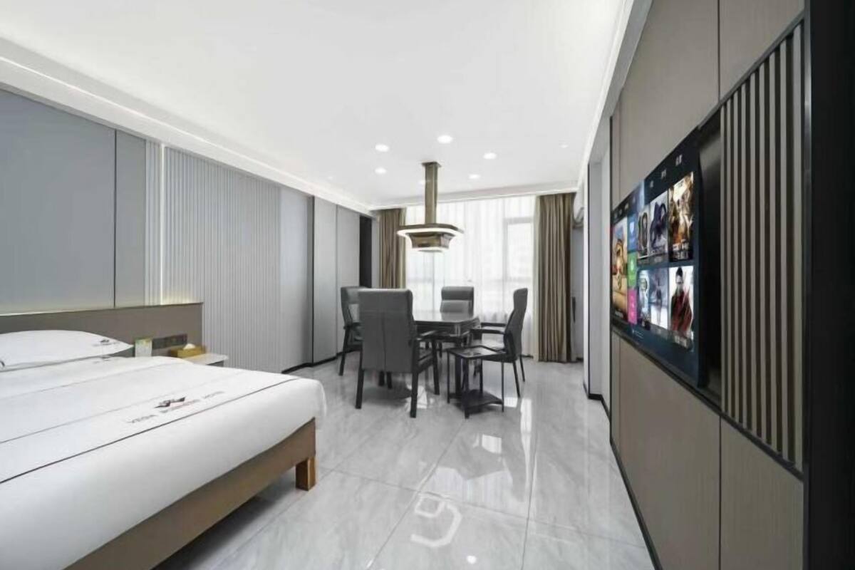 A modern hotel room with a bed, a dining table with chairs, a TV mounted on the wall, and a large window.
