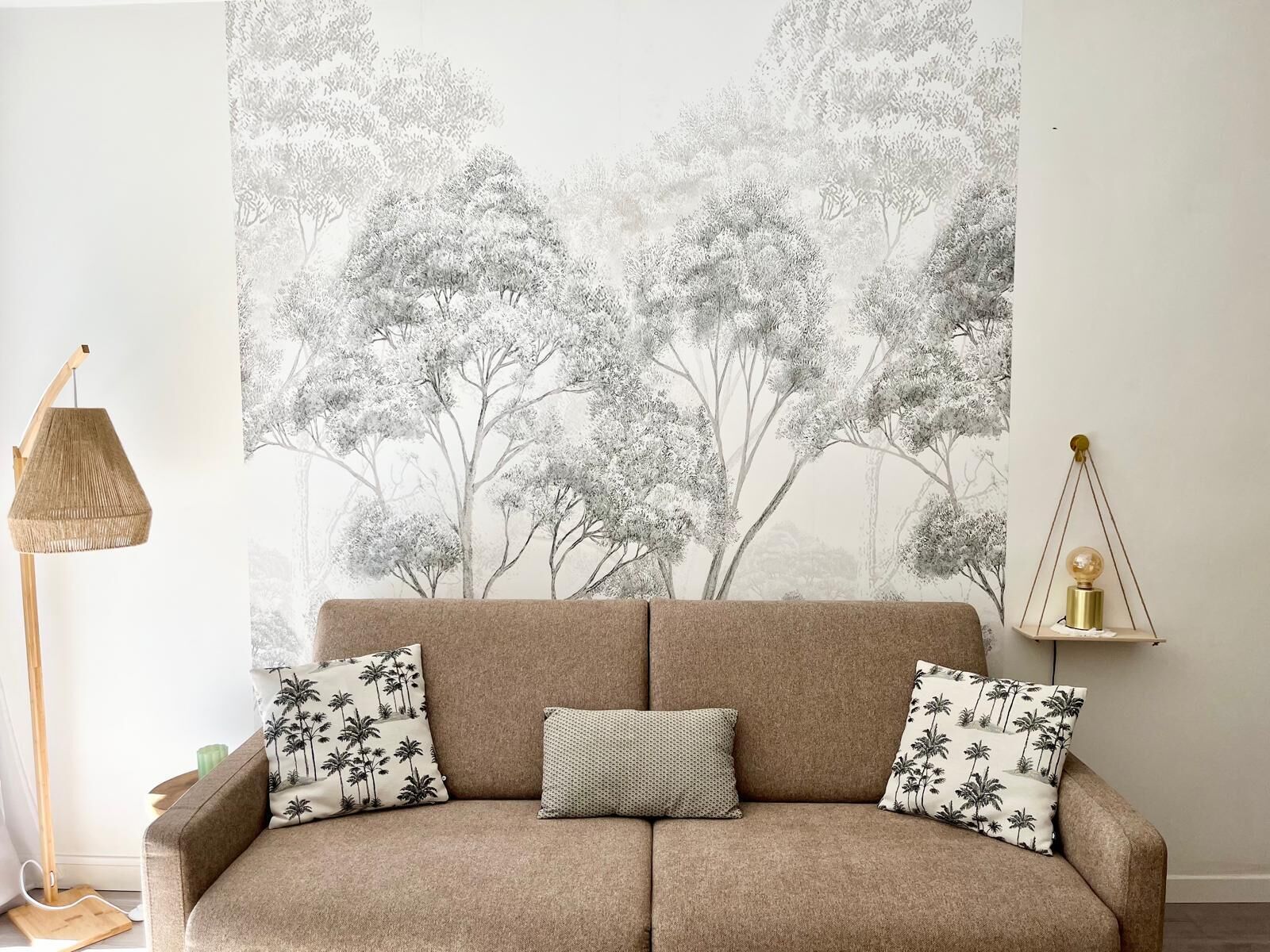 En beige sofa med tre puder, hvoraf den ene har et palmetræsmønster, i et rum med et stort tapet med et træmotiv på væggen.