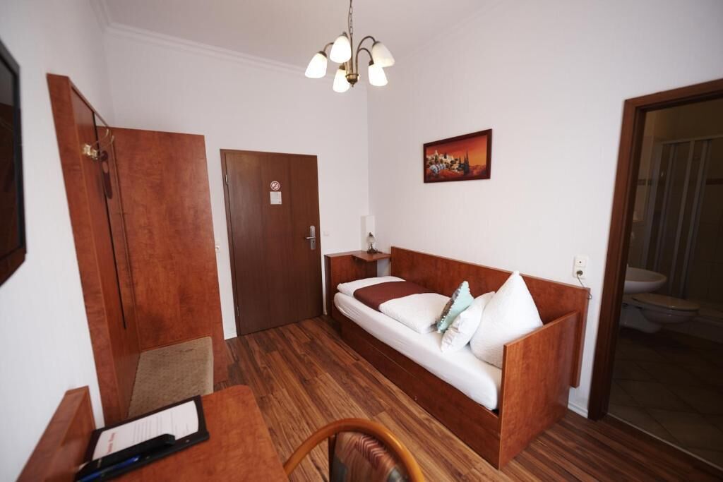 Una habitación con piso de madera, un sofá cama con almohadas, un escritorio con una silla, una puerta de madera, una lámpara de pared y un cuadro.