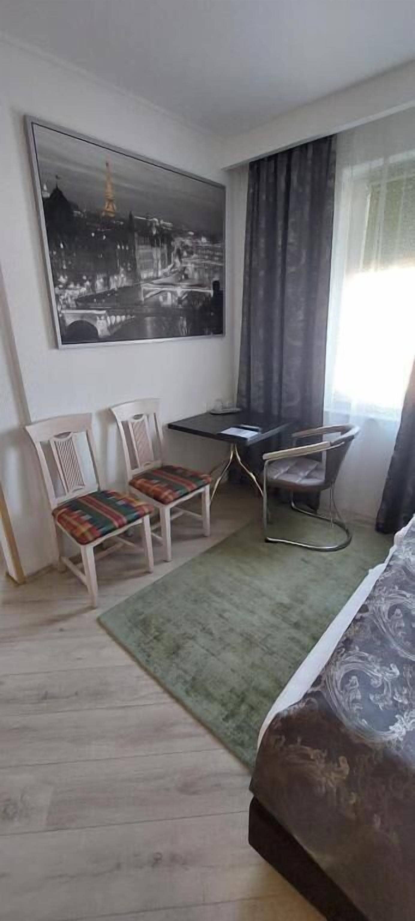 Ett hotellrum med en säng, ett skrivbord, två stolar, en matta och en stor målning med en stadsbild på väggen.