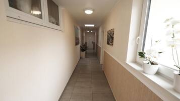 Un couloir étroit avec un sol carrelé, des murs blancs et une fenêtre ornée d’orchidées blanches.