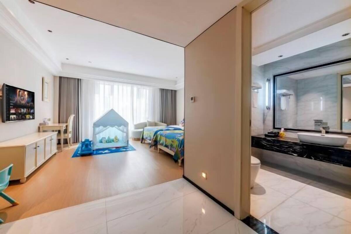 Een moderne hotelkamer met een bed, een bureau, een stoel, een televisie, een groot raam en een badkamer met een wastafel en spiegel.