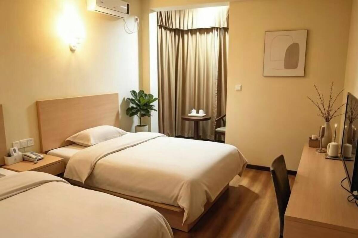 A hotel room with two beds, a desk, a chair, a plant, and a window with curtains.