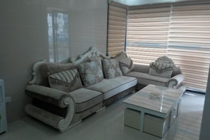 A modern living room with a sectional sofa, a coffee table, and a large window with blinds. - Haojia Express Hotel (Zhuhai Gongbei Port Fuhuali Branch) (Zhuhai)