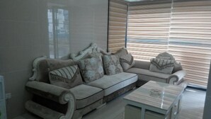 A modern living room with a beige sectional sofa, a glass coffee table, and a large window with blinds. - Haojia Express Hotel (Zhuhai Gongbei Port Fuhuali Branch) (Zhuhai)