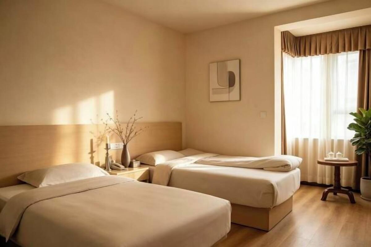 A hotel room with two beds, a bedside table, a small table with a plant, and a window with curtains.