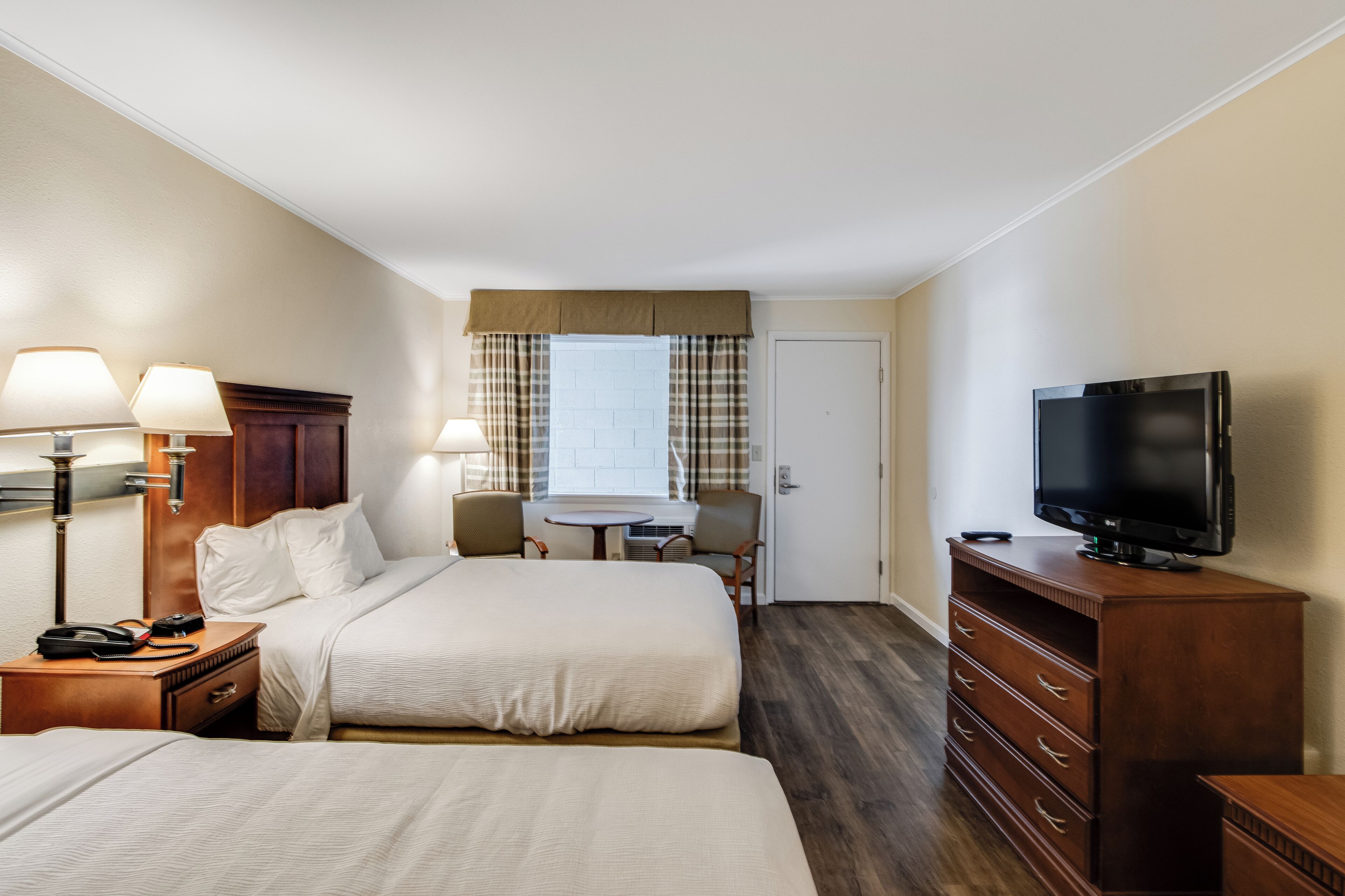 Ein Hotelzimmer mit zwei Betten, einem Fernseher, einem Schreibtisch mit Lampe, einem Sessel und einem Fenster mit Vorhängen.