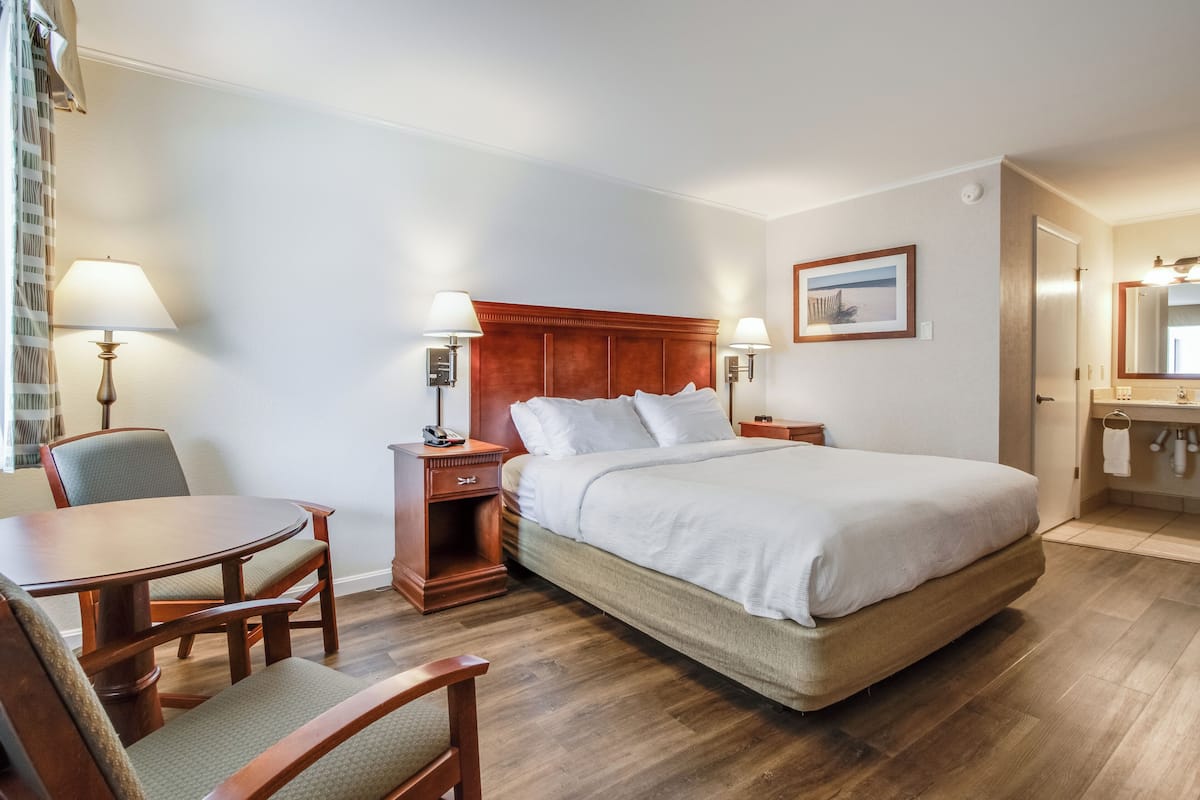 A hotel room with a large bed, a desk with two chairs, a small table, a nightstand, a bedside table, a mirror, and a framed picture on the wall.