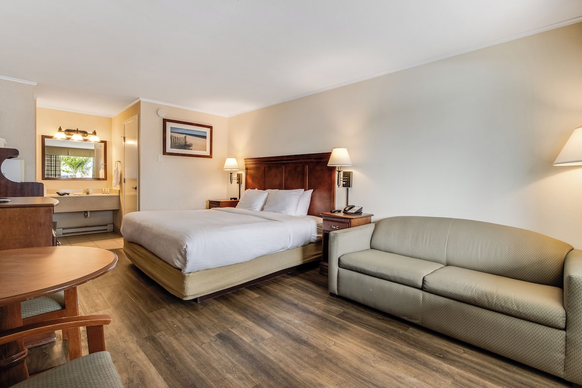 A hotel room with a bed, a sofa, a desk with a chair, a nightstand, and a lamp.