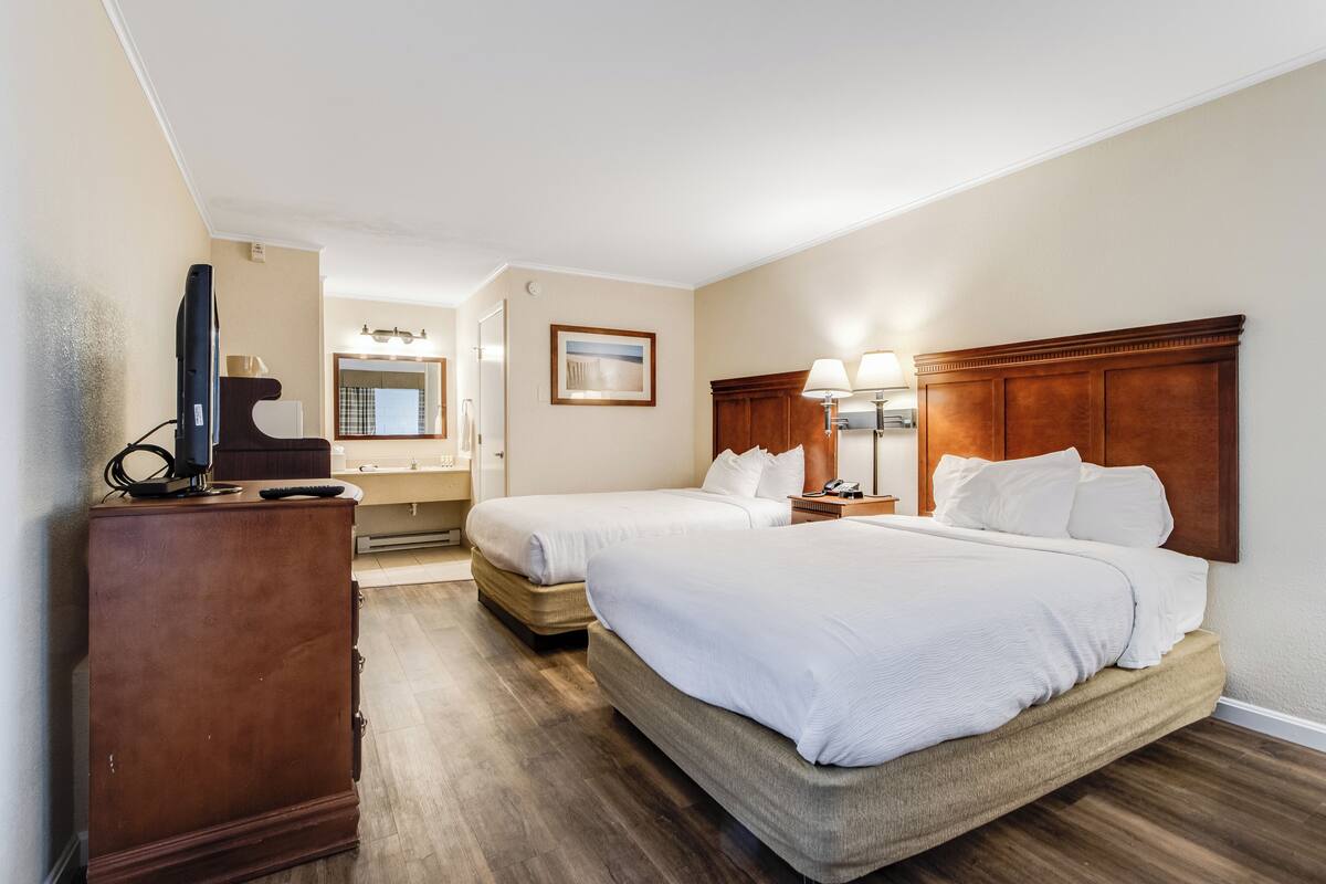 A hotel room with two beds, a dresser, a television, a mirror, and a bathroom visible in the background.