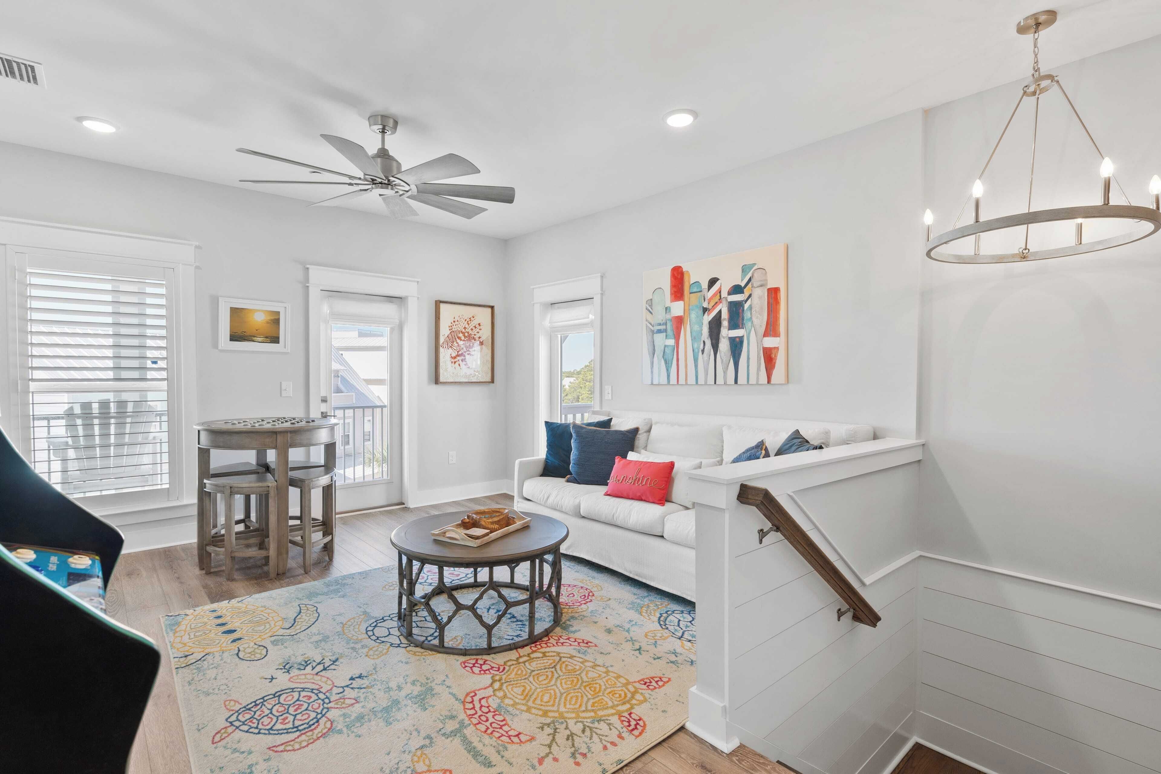 A spacious living room with a white sofa, a round coffee table, and a colorful rug. There is a ceiling fan, a chandelier, and a staircase with white railings.