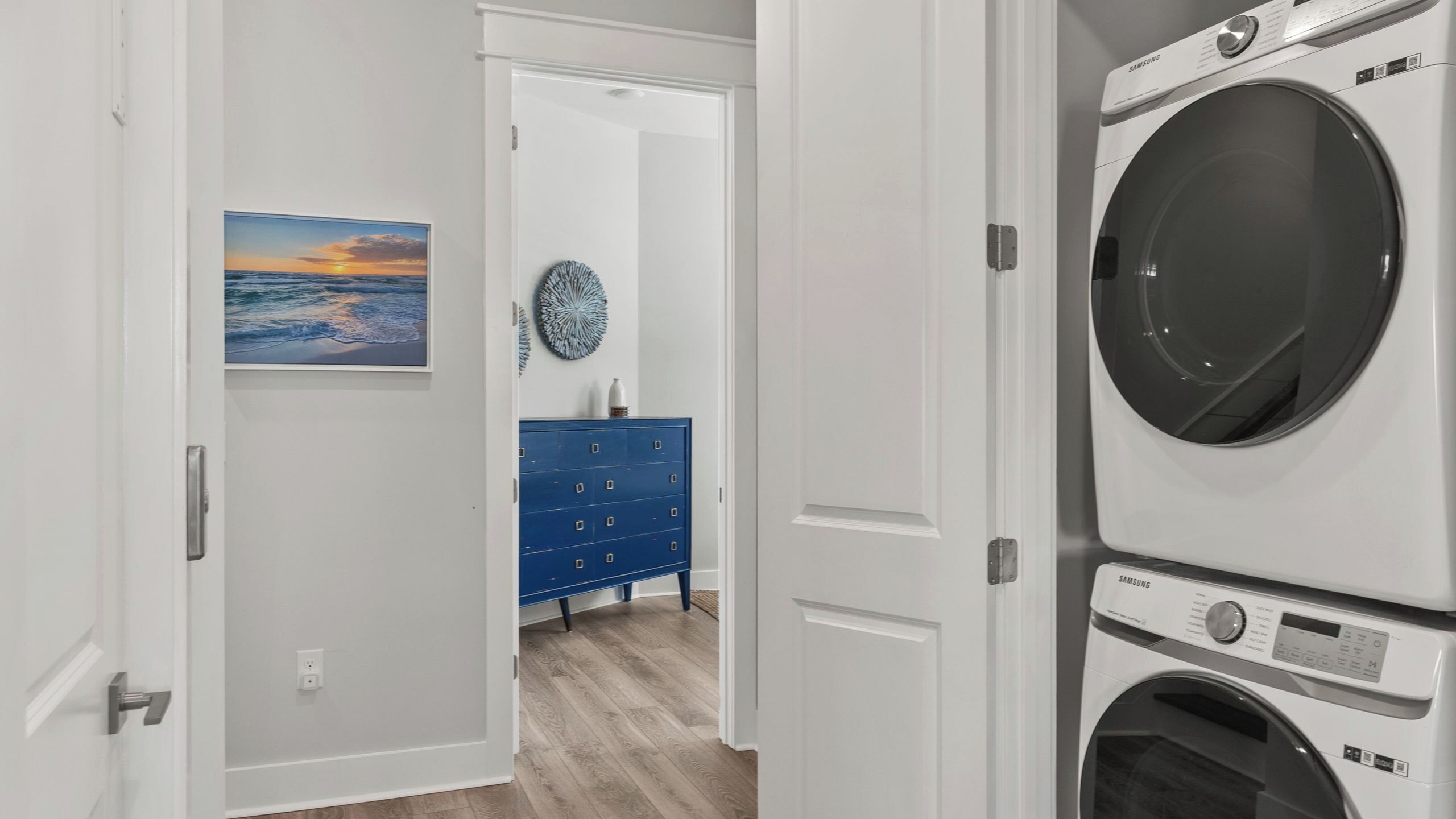 A laundry room with stacked washers and dryers, a blue dresser, a wall clock, and a painting of a sunset.