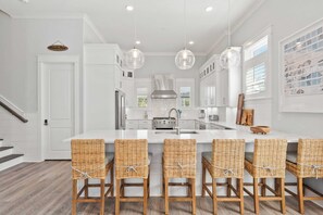 Cocina moderna con armarios blancos, una isla central con taburetes altos, luces colgantes y una campana extractora de acero inoxidable.