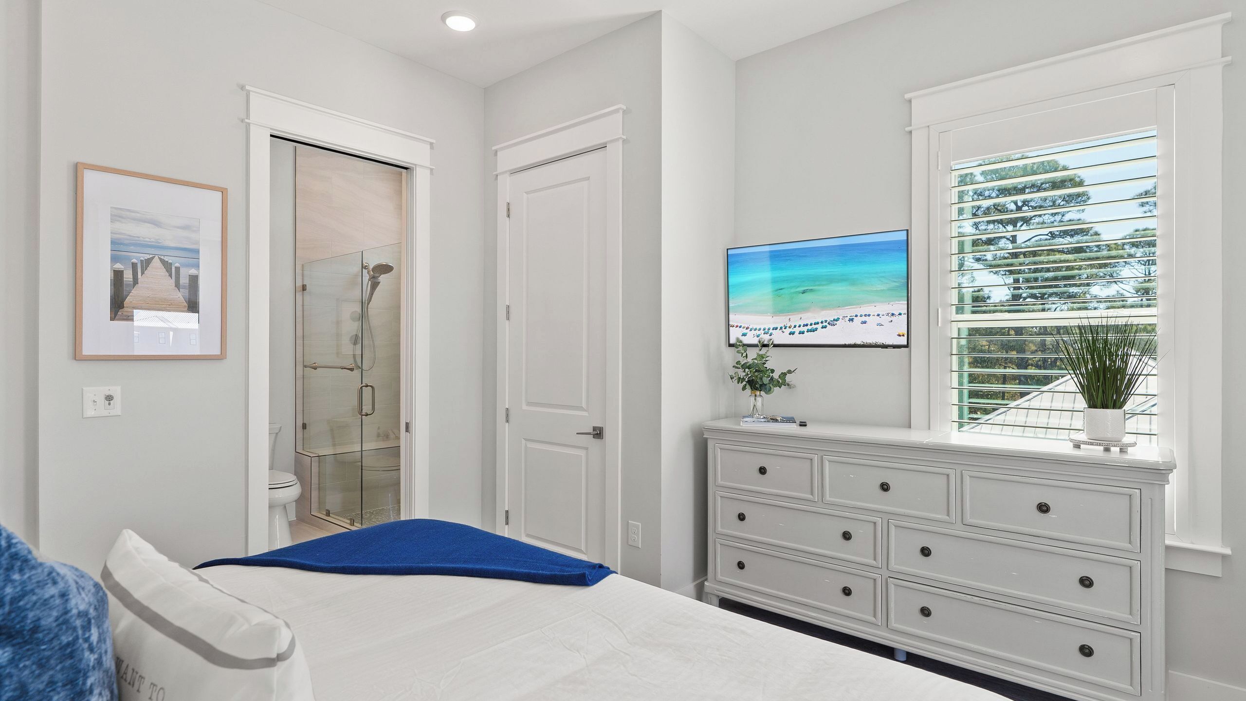 A bedroom with a bed, a dresser, a television, a window with blinds, and a shower.