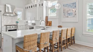 Une cuisine moderne avec des meubles blancs, un grand îlot central doté de tabourets, des luminaires suspendus et une hotte de cuisine en acier inoxydable.