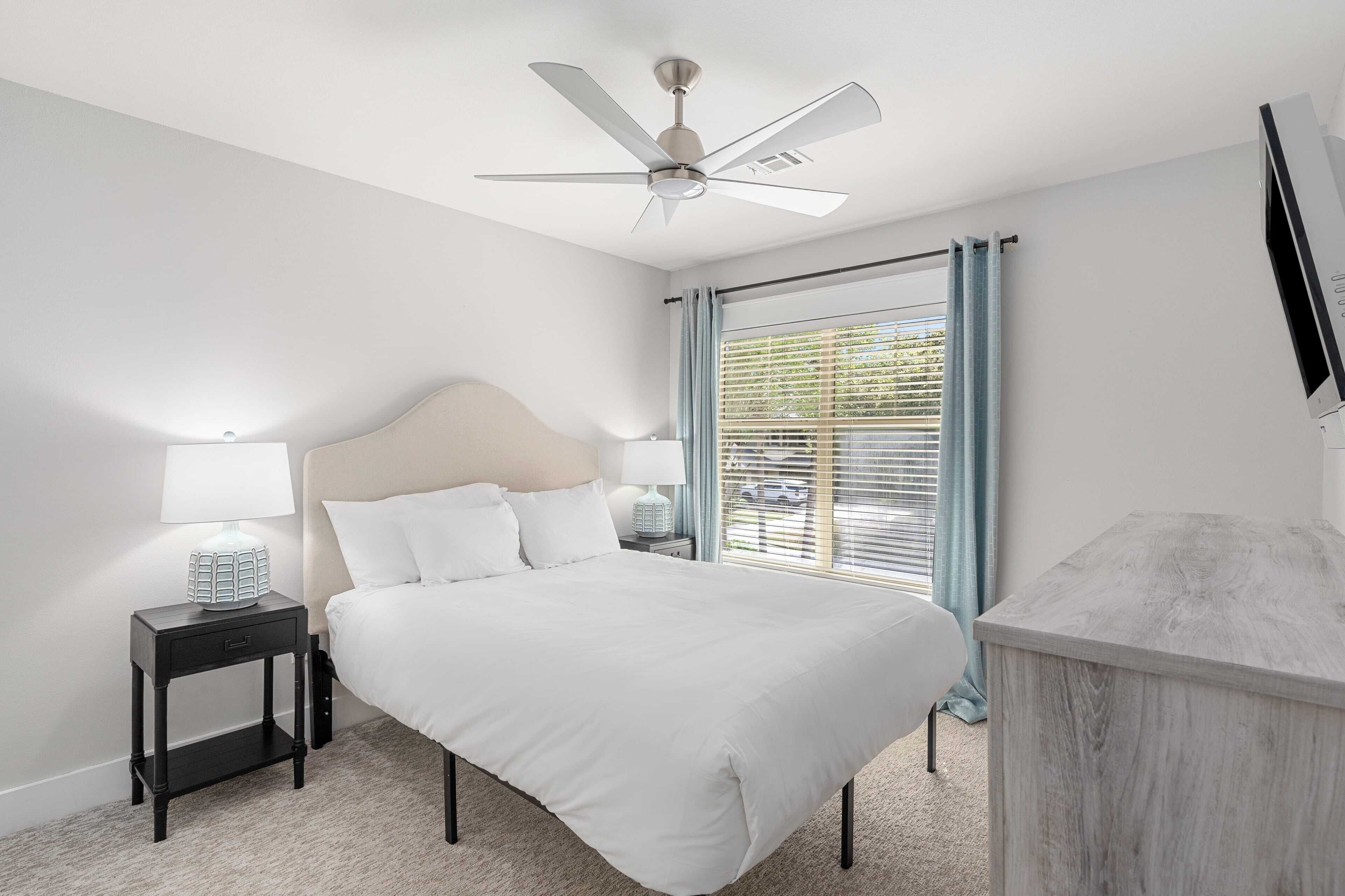 A bedroom with a bed, nightstands, a ceiling fan, a window with curtains, and a TV on the wall.