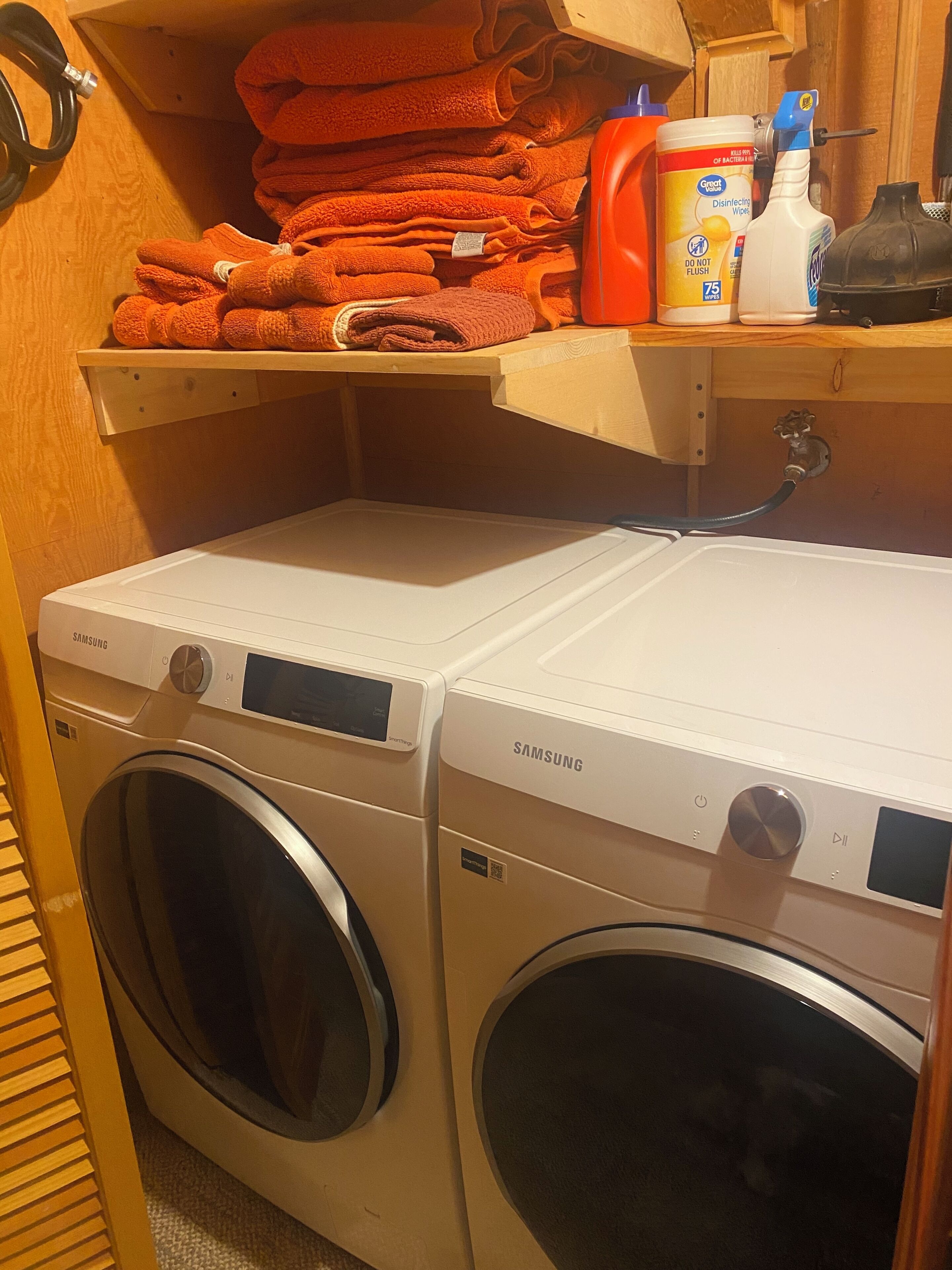A laundry area with two Samsung washing machines, a shelf with folded towels, and a bottle of disinfecting wipes.
