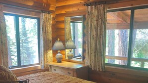 A wooden cabin bedroom with a bed, a desk, a lamp, and large windows.