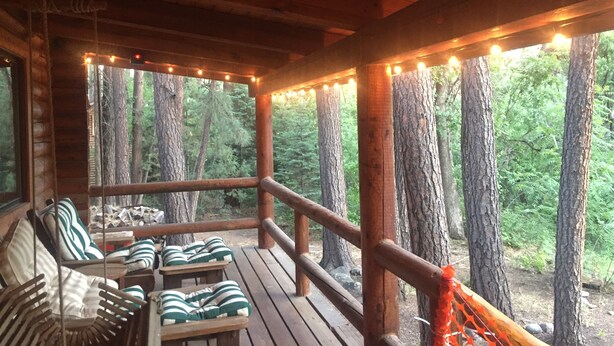 A wooden deck with string lights, wooden chairs with striped cushions, and a view of a forest.