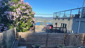 Eine Holzterrasse mit Gartenmöbeln, einem Maschendrahtzaun und Blick auf einen Parkplatz mit einem roten Fahrzeug.