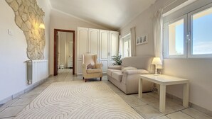 Ein modernes Wohnzimmer mit einer Wand aus Naturstein, einem beigen Sofa, einem kleinen Sessel, einem Couchtisch und Blick ins Freie durch ein großes Fenster.