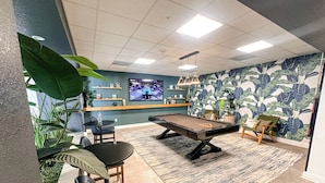 A pool table in a room with a blue wall, a large plant, and a television displaying a game. - Coastal Chic Apartment in St. Pete with Pool (St. Petersburg)