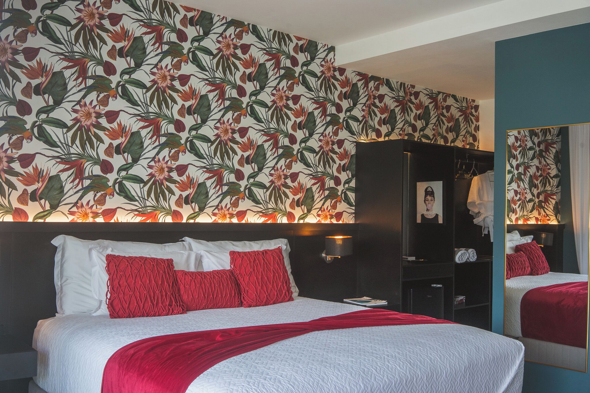 Een hotelkamer met een groot bed, rood-witte beddengoed, bloemenbehang, een kledingkast en een spiegel.
