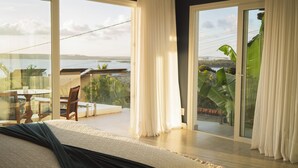 A bedroom with a large bed, a balcony with a table and chairs, and a view of a body of water and greenery. - The View Pipa (Tibau do Sul)