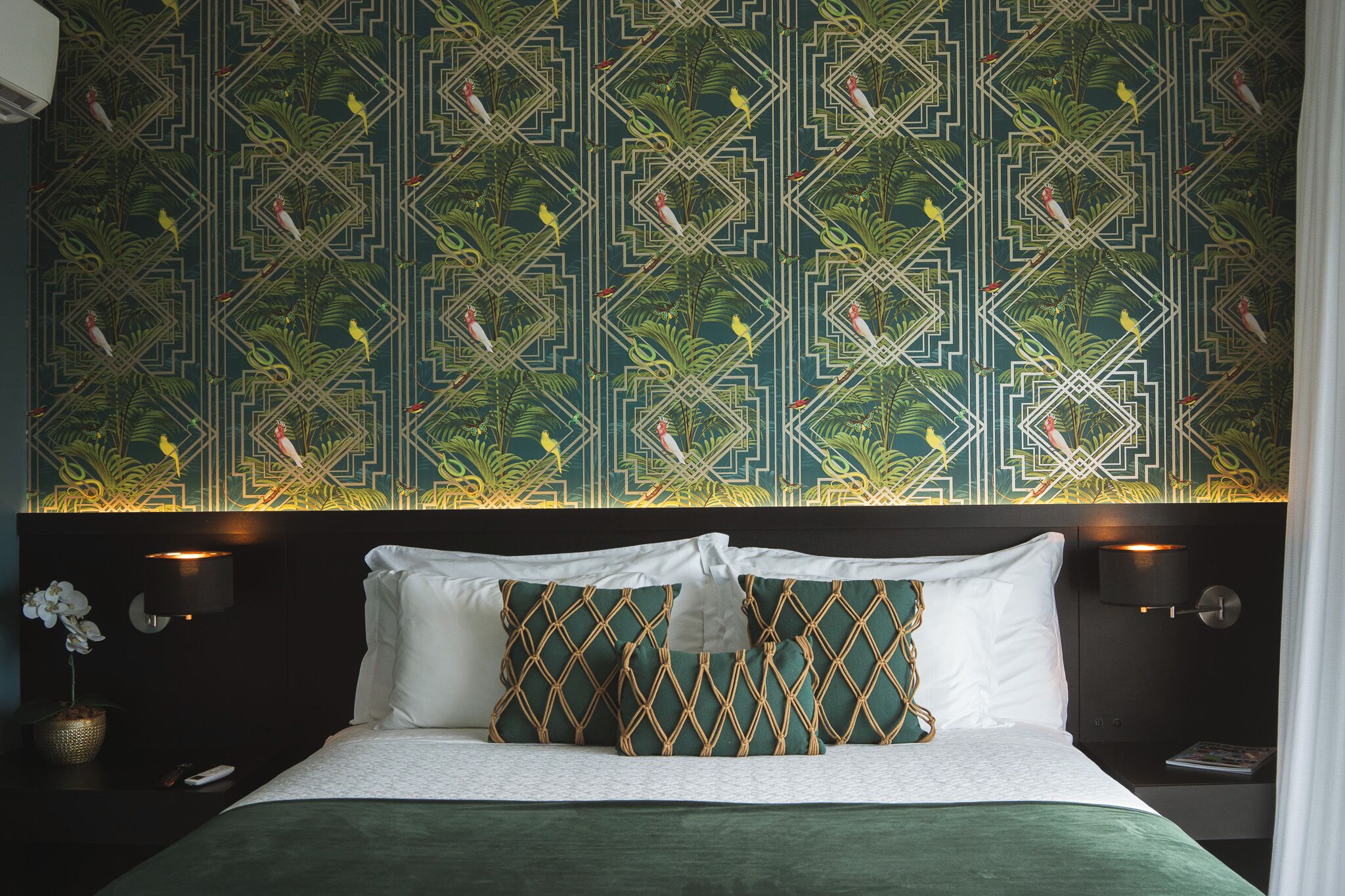A hotel room with a patterned wallpaper, a bed with white and green bedding, and two wall-mounted lamps.